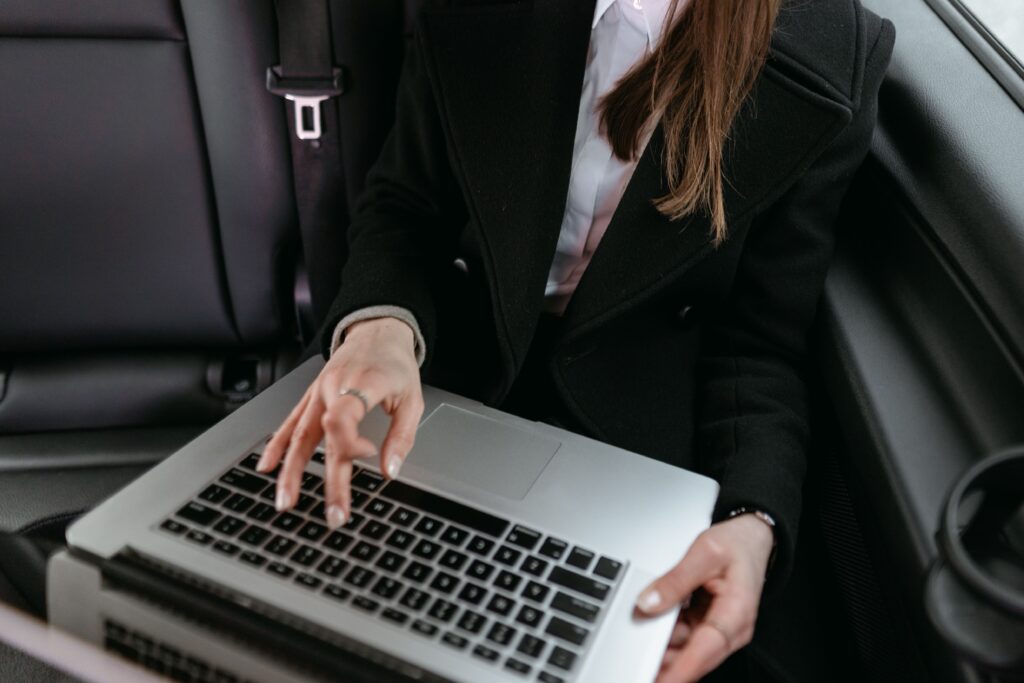 Person in a black coat typing on a silver laptop in the backseat of a car