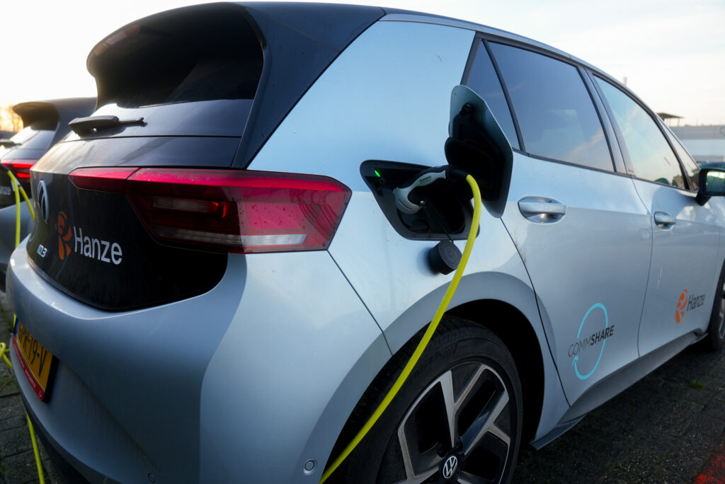 Blue electric SUV connected to a charging station with a yellow charging cable, rear and side doors visible, Hanze branding on the door.