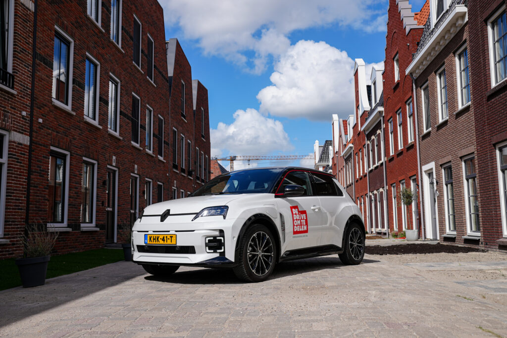 White SUV parked in a narrow brick street with red brick buildings on both sides and a blue sky with clouds above; car has a red Dutch license plate and a campaign decal on the door reading 'DE DRIVE OM TE DELEN'.